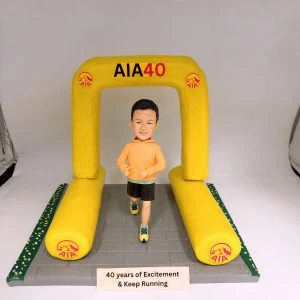 馬拉松主題人像公仔：短髮男子穿橙色連帽運動上衣、黑色短褲與黃藍跑鞋，在寫有「AIA40」的黃色充氣拱門下奔跑；拱門與兩側跑道邊緣印有 AIA 標誌，灰色路面前方放置銘牌「40 years of Excitement & Keep Running」。整體呈現AIA贊助的馬拉松比賽終點氛圍。