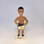 健身主題人像公仔：短髮男子公仔身穿粉紅條紋上衣與深灰長褲、白色運動鞋，雙手做出揮臂動作站在綠色草地造型底座上；旁邊放置黑藍色運動裝備袋與多件器材道具，前方木牌可見客製祝福字樣。背景為綠地與樹林場景的照片板，整體收納於透明展示盒內，呈現健身教練氣質的人像公仔擺設。