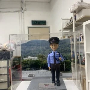 「香港警察學員人像公仔，身穿淺藍色長袖制服襯衫，佩戴黑色肩章與領帶，頭戴警帽，胸前名牌清晰可見，右手自然下垂，左手則做出敬禮般的姿勢，展現莊重與自信。公仔置於透明展示盒中，背景為綠意盎然的山景與訓練場地，象徵警隊訓練與守護社會的使命。底座附有字牌，內容提及『Intake 01/2023 警察學員結業禮』，可見此人像公仔為一份專屬訂製的結業紀念禮物，寓意學員順利完成培訓，邁向警務生涯的新起點。」