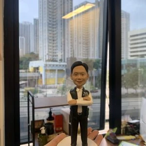 男性警察人像公仔，職級標示為總督察；短髮微笑，白襯衫配深色長褲與黑鞋，外穿深色戰術背心，胸前印有 POLICE 與臂章圖樣；雙臂交叉站姿。公仔立於白色圓形台座，前方金色名牌刻有祝賀升遷與贈送者資訊。背景為室內辦公環境與落地窗外的高樓城市景觀，由手掌托起近距離拍攝，呈現紀念與送禮氛圍。