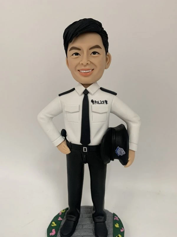 這是一份意義非凡的【晉升】賀禮，為一位年輕的【警務人員】訂製的專屬【人像公仔】。 從公仔的制服細節——尤其是肩上的一條銀色橫柴——我們可以推斷，這位【警務人員】榮升的職位是「見習督察」，這是其警隊生涯一個光輝的起點。公仔自信的笑容與輕鬆的站姿，正是這份喜悅的最佳寫照。 作品設計簡潔而專注，將所有焦點都集中在主角身上，底座的小花點綴，則象徵著他未來職業道路的無限生機。 這份禮物不僅僅是一件擺設，它更是一份認可、一個見證，紀念著他通過努力，成功邁入警官行列的決定性時刻。