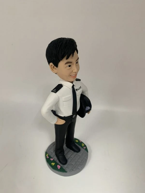 這是一份意義非凡的【晉升】賀禮，為一位年輕的【警務人員】訂製的專屬【人像公仔】。 從公仔的制服細節——尤其是肩上的一條銀色橫柴——我們可以推斷，這位【警務人員】榮升的職位是「見習督察」，這是其警隊生涯一個光輝的起點。公仔自信的笑容與輕鬆的站姿，正是這份喜悅的最佳寫照。 作品設計簡潔而專注，將所有焦點都集中在主角身上，底座的小花點綴，則象徵著他未來職業道路的無限生機。 這份禮物不僅僅是一件擺設，它更是一份認可、一個見證，紀念著他通過努力，成功邁入警官行列的決定性時刻。
