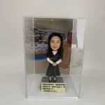 一款放置在透明亞加力膠盒中的客製化女士人像公仔，底座刻有聖經經文，背景為教室，是一份送給主日學老師或傳道人的感謝禮物。