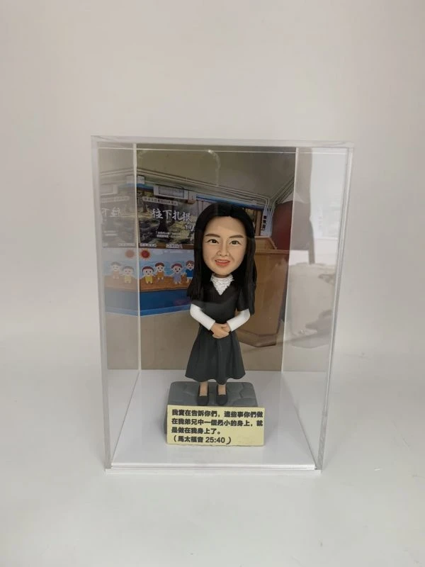 一款放置在透明亞加力膠盒中的客製化女士人像公仔，底座刻有聖經經文，背景為教室，是一份送給主日學老師或傳道人的感謝禮物。