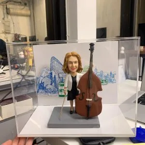 一款為女性低音大提琴演奏家訂製的人像公仔，公仔站在樂器旁，其背景配件裝飾為一張印有香港電車、渡輪及城市景觀的手繪風格圖畫。