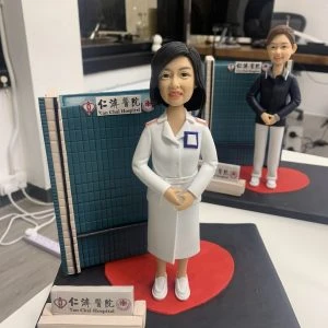 訂製仁濟醫院醫護人像公仔｜身穿白色醫師袍的醫生公仔站立於醫院屏風前，腳下有紅色心型底座，背景標示「仁濟醫院 Yan Chai Hospital」標誌。此公仔專為醫生與醫護人員打造，是獨特的專屬禮物與紀念擺設。
