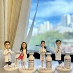 四位穿白袍的醫生／醫護人像公仔排成一列，背景為窗外藍天與高樓景致；每個公仔站在印有 JULAINE 字樣的底座旁，身側設有放大版「膠原增生注射」示範瓶模型，其中兩位手持托盤與疑似注射器，細節精緻，呈現醫學美容臨床感；整體作為醫生、醫護致謝或退休禮物極具紀念性。