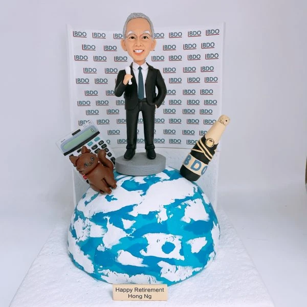 老闆退休主題的人像公仔：銀灰短髮、戴眼鏡的男士身穿黑色西裝與領帶，站在小型圓台上比出讚的手勢；前方為藍白地球造型底座，左側擺放大型計數機與棕色貓咪公仔，右側有綁著黑金緞帶的紅酒香檳瓶並貼有「BDO」字樣；背板重複排列「BDO」標誌，前方銘牌寫有「Happy Retirement」。整體呈現職場成就與個人喜好的退休禮物人像公仔佈置。