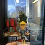 一個穿著馬拉松運動服、佩戴金色獎牌的人像公仔，站在綠色運動底座上，背景有聖誕樹和禮物堆。公仔穿著藍色T恤、黑色運動褲，戴著黃色運動帽，脖子掛著獎牌，底座上刻有「Great Boss Johnny, Happy Retirement!」的祝福語，以及Bausch+Lomb公司標誌。