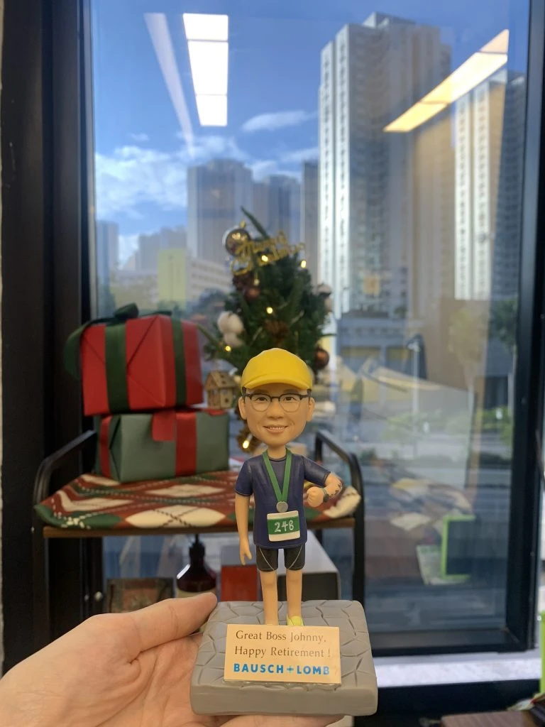 一個穿著馬拉松運動服、佩戴金色獎牌的人像公仔，站在綠色運動底座上，背景有聖誕樹和禮物堆。公仔穿著藍色T恤、黑色運動褲，戴著黃色運動帽，脖子掛著獎牌，底座上刻有「Great Boss Johnny, Happy Retirement!」的祝福語，以及Bausch+Lomb公司標誌。