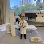 神經外科醫生人像公仔，身穿白袍，手持手術導管與醫療器材，背景擺放有大型醫療影像設備與病床，細緻還原醫護專業場景。此公仔為醫生、醫護人員專屬客製化禮物，適合升遷賀禮、退休紀念或醫護感謝之用。