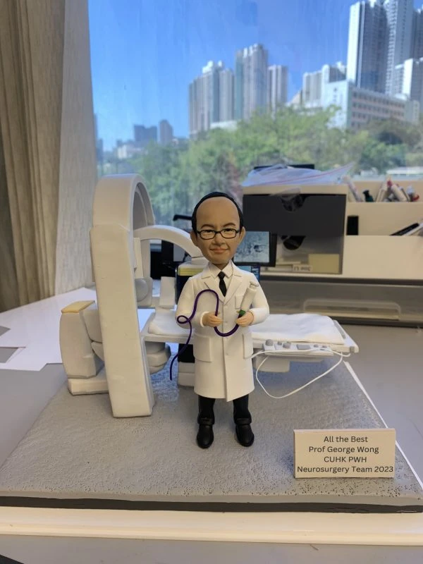 神經外科醫生人像公仔，身穿白袍，手持手術導管與醫療器材，背景擺放有大型醫療影像設備與病床，細緻還原醫護專業場景。此公仔為醫生、醫護人員專屬客製化禮物，適合升遷賀禮、退休紀念或醫護感謝之用。
