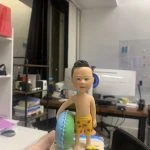 手掌上托著一尊兒童造型的游水人像公仔。公仔留短髮，赤裸上身，穿著黃色印有紅色小魚圖案的泳褲，右手自然下垂，左臂抱著綠藍雙色圓形泳圈。腳踩貝殼與細沙質感的底座，旁邊放著藍黃配色的小蛙鞋與小水池邊緣造型。Keywords：人像公仔，游水人像公仔。