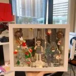 一個放在白色展示盒內的客製化人像公仔，場景以聖誕節為主題，周圍裝飾有聖誕老人、公仔雪人、聖誕樹、馴鹿與聖誕燈飾，背景營造溫馨的節日氛圍，非常適合作為聖誕節禮物或驚喜心意。