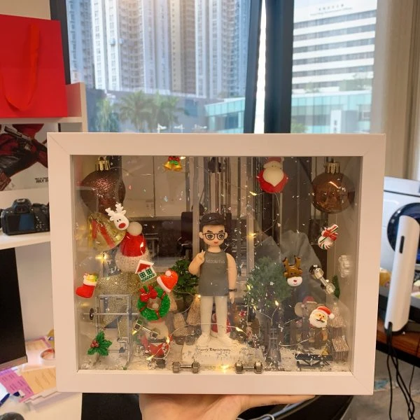 一個放在白色展示盒內的客製化人像公仔，場景以聖誕節為主題，周圍裝飾有聖誕老人、公仔雪人、聖誕樹、馴鹿與聖誕燈飾，背景營造溫馨的節日氛圍，非常適合作為聖誕節禮物或驚喜心意。