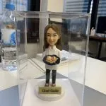 透明展示盒中的女廚師人像公仔：棕色長髮、白色廚師外套與黑色長褲，雙手端著深藍圓盤，上有捲餅與蔬菜擺盤；公仔立於白色圓形底座，前方金色名牌寫「Chef Gaile」。背景為辦公室窗景與桌面雜物，光線明亮，整體呈現典雅且具紀念性的女廚師退休禮物。