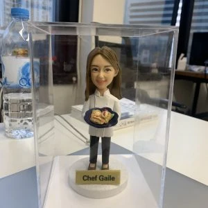 透明展示盒中的女廚師人像公仔：棕色長髮、白色廚師外套與黑色長褲，雙手端著深藍圓盤，上有捲餅與蔬菜擺盤；公仔立於白色圓形底座，前方金色名牌寫「Chef Gaile」。背景為辦公室窗景與桌面雜物，光線明亮，整體呈現典雅且具紀念性的女廚師退休禮物。