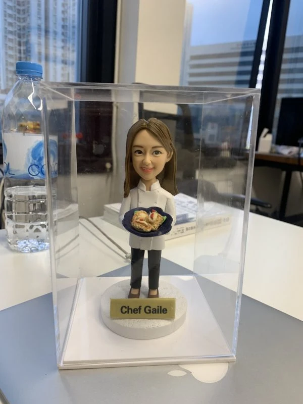 透明展示盒中的女廚師人像公仔：棕色長髮、白色廚師外套與黑色長褲，雙手端著深藍圓盤，上有捲餅與蔬菜擺盤；公仔立於白色圓形底座，前方金色名牌寫「Chef Gaile」。背景為辦公室窗景與桌面雜物，光線明亮，整體呈現典雅且具紀念性的女廚師退休禮物。