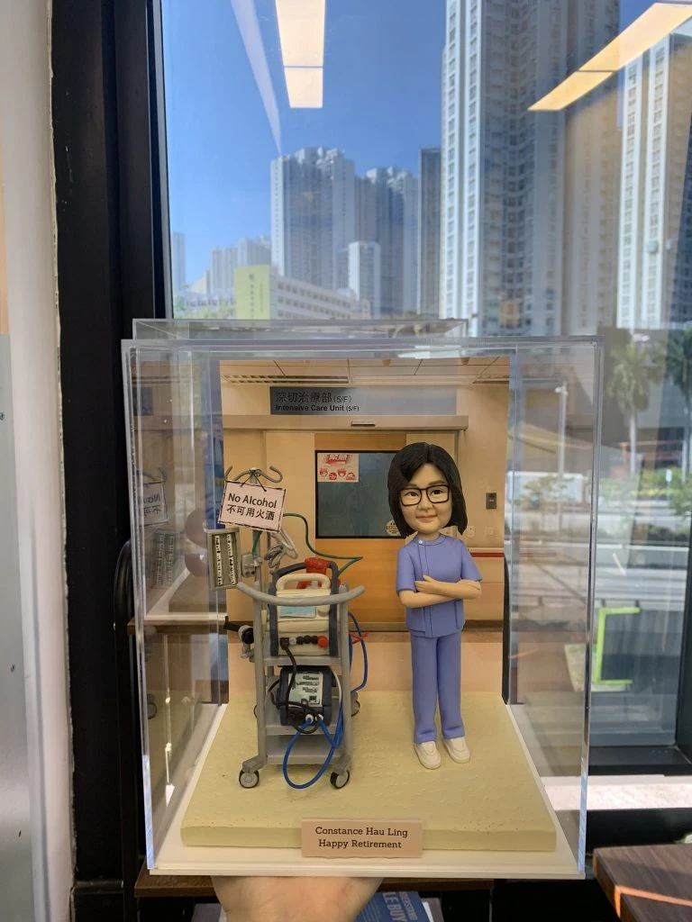 一款專屬定製的醫護人像公仔，身穿藍色護理制服，雙手交叉站立於醫療場景中。背景標示「深切治療部 (ICU)」，公仔旁邊擺放有醫療推車、監護設備與插管機，場景真實還原重症監護室環境。底座刻有「Constance Hau Ling Happy Retirement」字樣，作為醫生與醫護退休禮物極具紀念價值。