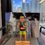 一個人像公仔，身穿馬拉松運動服裝，背號1103，站在模擬比賽場景中，做出勝利手勢。背景是一個城市景觀，展示了摩天大樓和樹木，底座上標有「Raymond Best Wishes for your New Chapter Nov 2024」的祝福語