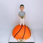 籃球教練主題人像公仔：笑容可掬的中年男子公仔，穿灰白格紋短袖襯衫與白色長褲、白鞋，手持金色感謝獎牌；他站在半圓形巨大橙色籃球造型底座上（黑色縫線清晰），前方貼有金色銘牌印上致敬與日期字樣。整體呈現向榮休籃球教練致意的儀式感場景，適合作為退休禮物的人像公仔。