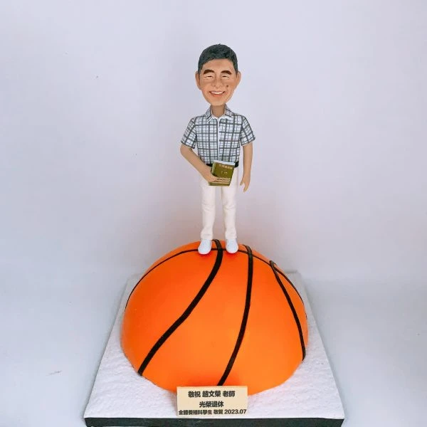 籃球教練主題人像公仔：笑容可掬的中年男子公仔，穿灰白格紋短袖襯衫與白色長褲、白鞋，手持金色感謝獎牌；他站在半圓形巨大橙色籃球造型底座上（黑色縫線清晰），前方貼有金色銘牌印上致敬與日期字樣。整體呈現向榮休籃球教練致意的儀式感場景，適合作為退休禮物的人像公仔。
