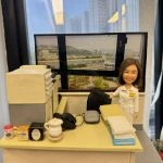 透明展示盒內的辦公室場景人像公仔：一位短髮女性穿白色制服、肩章與名牌，站在米黃色書桌旁微笑；桌上有文件疊、座機電話、滑鼠、茶具與幾罐沖泡品，左側是一部多功能影印／掃描器；背後窗景為臨河城市與山景的大幅相片，呈現醫院或醫療機構行政辦公風格。