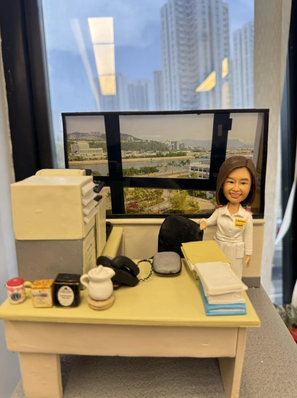 透明展示盒內的辦公室場景人像公仔：一位短髮女性穿白色制服、肩章與名牌，站在米黃色書桌旁微笑；桌上有文件疊、座機電話、滑鼠、茶具與幾罐沖泡品，左側是一部多功能影印／掃描器；背後窗景為臨河城市與山景的大幅相片，呈現醫院或醫療機構行政辦公風格。