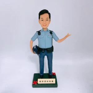 一尊以「警員」職級為主題的人像公仔置於白色無縫背景前拍攝。公仔黑色短髮、微笑神情；身穿淺藍色短袖制服襯衫，肩上有深色肩墊與扣件，左胸口袋上方印有 POLICE 字樣。腰間繫黑色裝備腰帶，配有多個裝備袋與扣具；下身為深藍長褲與黑色皮鞋。右手自然下垂並持一頂深藍色頭盔，左臂抬起向側前方打開手掌做示意/招呼姿態。腳下為深綠色方形台座，前側裝有米金色名牌，刻有「爸爸退休快樂」等祝福字樣，台座兩角點綴紅色小花。整體以簡潔白底呈現，凸顯公仔與制服細節，營造紀念與致意的送禮場景。