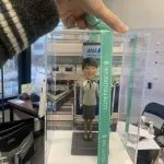一款為全日空（ANA）航空女地勤人員訂製的人像公仔，公仔身穿制服，被放置在一個透明禮盒中，其背景為機場櫃檯的配件裝飾。