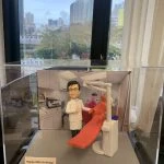 一座放在透明展示盒內的牙科主題人像公仔：公仔身穿白色醫生袍與眼鏡，右手比讚，站在紅色牙科治療椅旁；椅側配有牙科器械柱、口腔吸引系統與控制面板，頭頂延伸手臂式無影燈；背景為診療室櫥櫃與儀器照片。底座前方小牌寫有「Happy 60th Birthday!」，呈現為牙科醫護人員的客製紀念禮物。