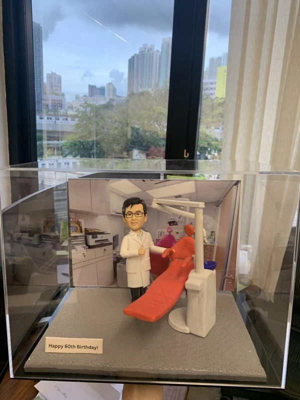 一座放在透明展示盒內的牙科主題人像公仔：公仔身穿白色醫生袍與眼鏡，右手比讚，站在紅色牙科治療椅旁；椅側配有牙科器械柱、口腔吸引系統與控制面板，頭頂延伸手臂式無影燈；背景為診療室櫥櫃與儀器照片。底座前方小牌寫有「Happy 60th Birthday!」，呈現為牙科醫護人員的客製紀念禮物。