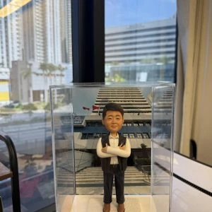 客製化警察人像公仔，身穿白襯衫與黑色背心，雙手交叉微笑站立，底座配有專屬銘牌，背景為警署大樓。銘牌刻有「偵緝警察 陳俊豪 祝一帆風順 步步高陞 荃灣刑事調查隊第三隊全人」字樣，象徵同袍對升職的祝賀與鼓勵。這款警察人像公仔適合用於升職賀禮、榮休紀念或作為警務人員的專屬禮物收藏。