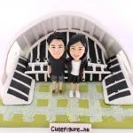 一款情侶人像公仔，一男一女身穿黑色情侶裝，親密地站立在一個微縮的足球觀衆席場景前，象徵他們對運動的共同熱愛。