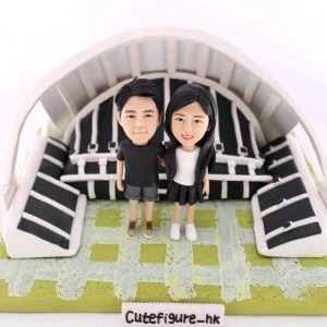 一款情侶人像公仔，一男一女身穿黑色情侶裝，親密地站立在一個微縮的足球觀衆席場景前，象徵他們對運動的共同熱愛。