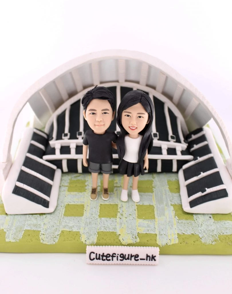 一款情侶人像公仔，一男一女身穿黑色情侶裝，親密地站立在一個微縮的足球觀衆席場景前，象徵他們對運動的共同熱愛。