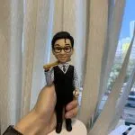 一款棒球主題的人像公仔：短黑髮、戴黑框眼鏡的男子露出微笑，穿著藍白直條襯衫、黑色針織背心與西裝長褲，左手握有棒球，右肩自然扛著木質球棒；腳踏黑色皮鞋，站在白色圓形底座上。背景為室內窗簾與城市景色的淺景深，前景可見手掌托起公仔。此作品強調職場紳士風與運動元素結合的棒球人像公仔與人像公仔。