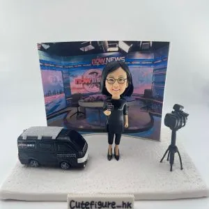 新聞採訪主題人像公仔場景：短髮戴眼鏡的女性記者穿黑色連身裙與高跟鞋，手持麥克風站在白色地台上；左側是一台黑色新聞採訪車模型，右側為架在三腳架上的專業攝影機；背板呈現演播室畫面與節目佈景。整體具專業與儀式感，適合作為新聞記者退休禮物與致敬紀念擺設。