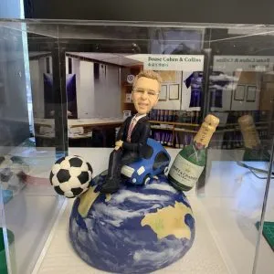 外國老闆主題的人像公仔坐在藍色小車上,整體擺設置於藍白相間的地球造型球體上;公仔穿深色西裝、紅黑條紋領帶與黑皮鞋,面帶微笑、配戴眼鏡。地球一側擺有黑白相間的足球,另一側立著香檳酒瓶(MOËT & CHANDON)造型,背景板為辦公室書架與公司字樣「Boase Cohen & Collins」。底座為白色紋理平台並置於透明展示盒中,呈現外國老闆退休禮物的人像公仔作品。