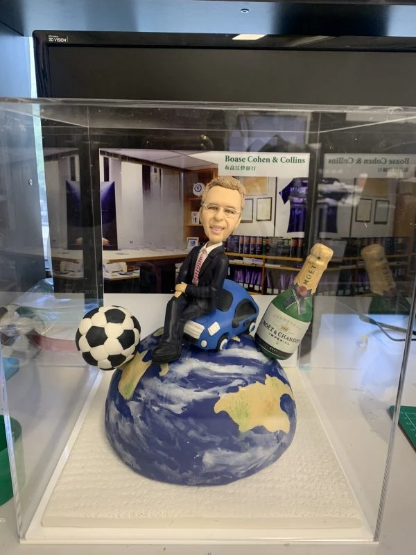 外國老闆主題的人像公仔坐在藍色小車上，整體擺設置於藍白相間的地球造型球體上；公仔穿深色西裝、紅黑條紋領帶與黑皮鞋，面帶微笑、配戴眼鏡。地球一側擺有黑白相間的足球，另一側立著香檳酒瓶（MOËT & CHANDON）造型，背景板為辦公室書架與公司字樣「Boase Cohen & Collins」。底座為白色紋理平台並置於透明展示盒中，呈現外國老闆退休禮物的人像公仔作品。