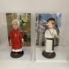 兩款並排展示在透明亞加力膠盒中的客製化神父人像公仔，左邊身穿紅色祭披，右邊身穿白色聖袍比讚，是送給牧師或神職人員的禮物系列。