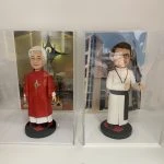 兩款並排展示在透明亞加力膠盒中的客製化神父人像公仔，左邊身穿紅色祭披，右邊身穿白色聖袍比讚，是送給牧師或神職人員的禮物系列。