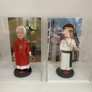 兩款並排展示在透明亞加力膠盒中的客製化神父人像公仔,左邊身穿紅色祭披,右邊身穿白色聖袍比讚,是送給牧師或神職人員的禮物系列。
