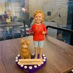 女同事退休人像公仔，身穿紅色上衣與淺藍色褲子，手持一隻紫色運動鞋，身旁有一隻可愛的橘色貓咪。公仔站立於紫色心形底座上，底座上有白色小裝飾並附上祝賀退休的字牌，作為特別的退休禮物。