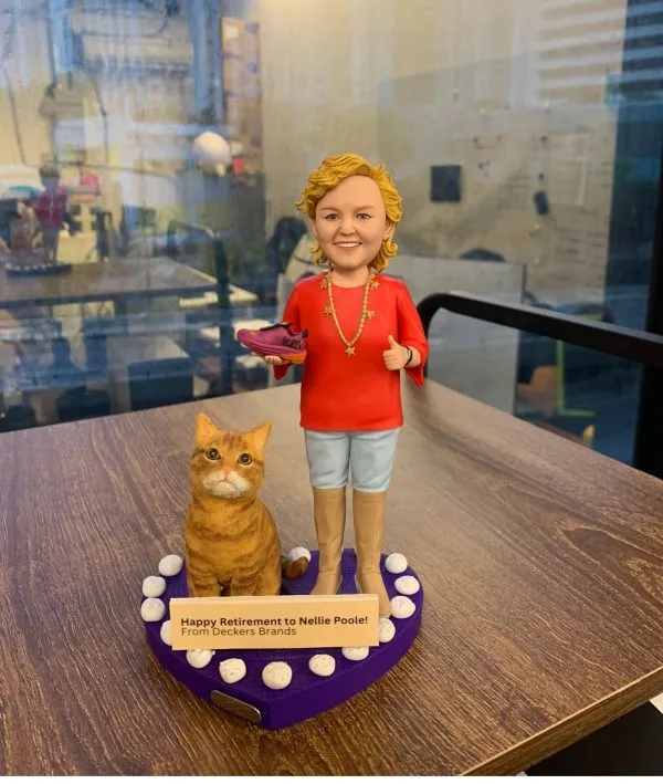 女同事退休人像公仔，身穿紅色上衣與淺藍色褲子，手持一隻紫色運動鞋，身旁有一隻可愛的橘色貓咪。公仔站立於紫色心形底座上，底座上有白色小裝飾並附上祝賀退休的字牌，作為特別的退休禮物。