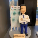 一款為Nittan Capital老闆訂製的退休禮物。精緻的人像公仔雙手抱胸，自信地站在公司大樓模型旁邊，底座銘牌刻有「Happy retirement September 2024」，是一份極具心思的公司禮物，彰顯了對領導者的敬意。
