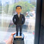 一款大學教授的人像公仔，他戴著眼鏡，身穿黑色西裝，站立在一個黑色的獎盃底座上，底座刻有他的姓名「Lawrence Lau」以及所屬機構「PolyU CFSO DoCFS」。