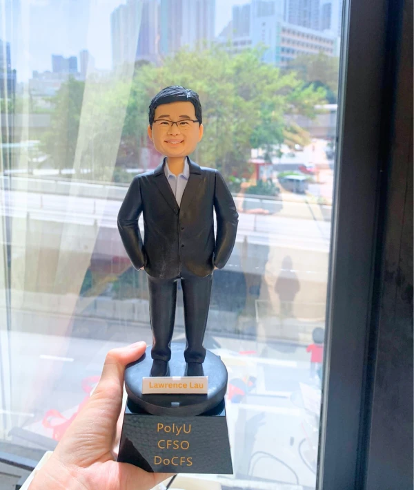 一款大學教授的人像公仔，他戴著眼鏡，身穿黑色西裝，站立在一個黑色的獎盃底座上，底座刻有他的姓名「Lawrence Lau」以及所屬機構「PolyU CFSO DoCFS」。