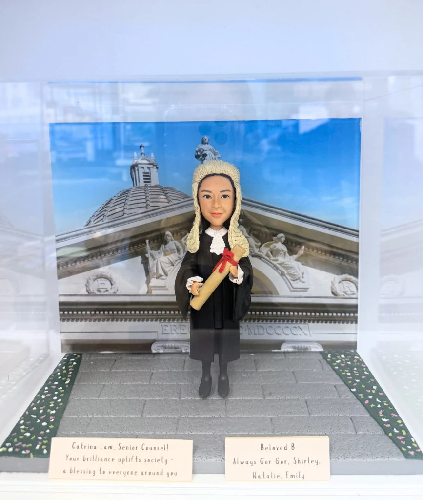 一款女性資深大律師人像公仔，身穿律師袍、頭戴假髮，站立在香港終審法院背景前，是慶祝法律畢業或晉升的完美祝賀禮物及畢業禮物。