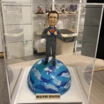 透明展示盒內的人像公仔：短髮戴眼鏡的男士穿深灰西裝，雙手做掀開西裝的動作，露出內層藍色上衣與紅黃色「10」字樣徽章，象徵十周年慶。公仔站在藍白相間的地球模型上，底座前方貼有金色標牌，刻有「精益求精 領先同儕」字樣。背景為工作室環境與層架擺設。整體呈現專業且富儀式感的公司周年禮物擺設。